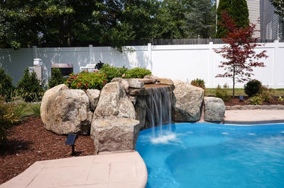 Celilo C - Evesham, NJ - This custom installation features a multi-tier natural stone waterfall cascading into a gunite pool. The waterfall is constructed from large, irregularly shaped boulders arranged to create a natural, freeform cascade that flows into the pool below. The surrounding area is landscaped with mulch, shrubs, and a small Japanese maple tree, enhancing the serene backyard setting.