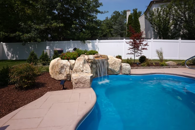 Celilo D - Evesham, NJ - This custom installation features a multi-tier natural stone waterfall cascading gently into a gunite pool, accented by large river boulders and integrated landscaping. The waterfall includes a tiered spillway design that creates a soothing water flow, while the surrounding area is landscaped with mulch, shrubs, and a low-maintenance white vinyl fence. The project demonstrates high-quality craftsmanship with seamless integration of natural materials and a clean, inviting poolside environment.