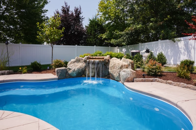 Celilo E - Evesham, NJ - This professionally installed outdoor feature showcases a natural stone waterfall cascading from a tiered rock formation into a pristine gunite pool. The waterfall, constructed with large, irregularly shaped boulders, creates a soothing cascade that flows seamlessly into the pool's clear blue water. The surrounding area is meticulously landscaped with shrubs, small trees, and a neatly maintained white privacy fence, enhancing the tranquil backyard setting in Evesham, NJ.
