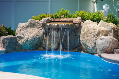Celilo F - Evesham, NJ - This custom installation features a multi-tier natural stone waterfall cascading into a gunite pool, surrounded by large boulders that enhance the natural aesthetic. The waterfall includes a tiered spillway with smooth, flowing water creating a soothing visual and auditory experience. The surrounding landscaping includes vibrant flowering shrubs, contributing to a serene backyard setting.