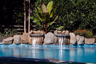 Cimahi A - Medford, NJ - This custom installation features a natural stone waterfall with multiple cascading tiers, seamlessly integrated into a gunite pool. The waterfall is constructed from large, irregularly shaped boulders that create a rustic, organic look, while the pool itself has a smooth, turquoise finish. Surrounded by dense tropical foliage and vibrant flowering plants, the setting enhances the tranquil and luxurious ambiance of the outdoor living space in Medford, NJ.