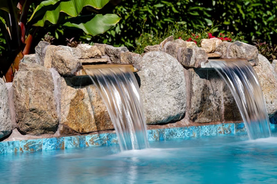 Cimahi C - Medford, NJ - This custom installation features a multi-tier natural stone waterfall cascading gracefully into a gunite pool. The waterfall is constructed with large, irregularly shaped boulders that create a rugged, organic look, while the pool edge is finished with a blue mosaic tile accent that contrasts beautifully with the earthy tones of the stone. The setting is a well-maintained backyard with lush greenery and tropical plants in the background, enhancing the serene and luxurious atmosphere of the outdoor living space.