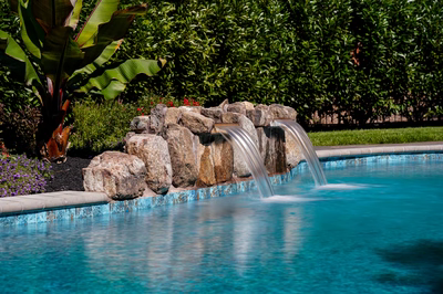 Cimahi D - Medford, NJ - This custom installation features a natural stone waterfall cascading from a multi-tiered rock formation into a gunite pool. The waterfall includes a tiered cascade with smooth, polished stone surfaces that guide the water flow, creating a soothing visual and auditory experience. The surrounding area is landscaped with lush greenery, including banana plants and flowering shrubs, enhancing the natural aesthetic of the outdoor living space in Medford, NJ.