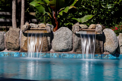 Cimahi F - Medford, NJ - This custom installation features a multi-tier natural stone waterfall cascading into a gunite pool with blue tile accents. The waterfall is constructed from large, irregularly shaped boulders arranged to create a natural, freeform cascade that flows into the pool below. The surrounding area is landscaped with lush tropical foliage, including banana plants, enhancing the serene and luxurious backyard setting.