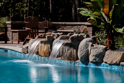 Cimahi G - Medford, NJ - This custom installation features a natural stone waterfall with two cascading streams flowing over large river rocks into a gunite pool. The waterfall is integrated with a tiered stone grotto, showcasing expert craftsmanship through precise stone placement and seamless water flow. The surrounding area includes a stone deck, wooden lounge chairs, and lush tropical landscaping, creating an inviting backyard oasis.