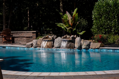 Cimahi H - Medford, NJ - This custom installation features a natural stone waterfall cascading from large boulders into a gunite pool with a blue tile coping. The waterfall includes multiple tiers with smooth, flowing water, creating a tranquil water feature. The surrounding area is landscaped with tropical plants and a brick wall, enhancing the serene backyard setting.