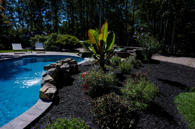 Cimahi J - Medford, NJ - This custom installation features a natural stone waterfall cascading from tiered boulders into a gunite pool with a smooth, curved edge. The waterfall is integrated seamlessly with the surrounding landscaping, using dark mulch beds and vibrant flowering plants to create a tranquil backyard oasis. The construction demonstrates high-quality craftsmanship with precise stone placement and a flowing water feature that enhances the serene atmosphere of the outdoor living space.