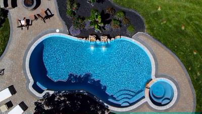 Cimahi L - Medford, NJ - This professionally installed gunite pool features a prominent natural stone waterfall cascading from a raised tier into the main pool area. The waterfall is integrated with carefully placed boulders that create a natural, flowing appearance. The pool itself has clean lines and a smooth finish, surrounded by a well-maintained brick patio and lush landscaping, creating an inviting outdoor living space.