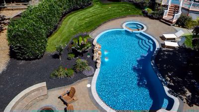 Cimahi M - Medford, NJ - This professional installation features a kidney-shaped gunite pool with a natural stone waterfall cascading into the main swimming area. The pool showcases clean lines and excellent construction quality with smooth concrete coping and integrated stone accents. The surrounding landscape includes manicured grass, strategically placed shrubs and flowering plants, and comfortable wooden Adirondack chairs creating an inviting outdoor living space.