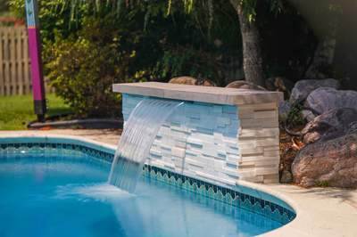 Corona C - Mt. Laurel, NJ - This professional installation features a sleek, modern waterfall constructed from light blue tiles with a textured finish, cascading smoothly into a gunite swimming pool. The waterfall is integrated into a custom concrete structure with a flat, tiled coping and decorative scalloped edge along the pool. The surrounding area includes natural boulders and lush greenery, creating a serene backyard oasis in Mt. Laurel, NJ.