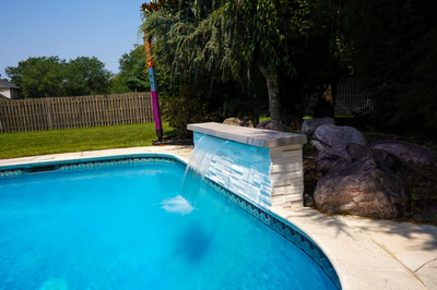 Corona D - Mt. Laurel, NJ - This professional installation features a sleek, modern waterfall cascading from a concrete and tile spillway into a pristine gunite infinity pool. The waterfall is accented with natural stone boulders and integrated seamlessly into the pool's edge, creating a clean, contemporary look. The surrounding backyard includes well-maintained grass, mature trees providing shade, and a wooden fence for privacy, enhancing the luxurious outdoor living space.