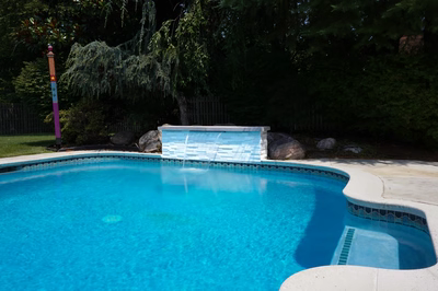 Corona E - Mt. Laurel, NJ - This professional installation features a custom-built stone waterfall with multiple tiers cascading into a gunite swimming pool. The waterfall is constructed from natural stone blocks, creating a clean, modern aesthetic that contrasts beautifully with the surrounding lush greenery. The pool itself has a smooth, reflective blue surface and is bordered by a decorative tile edge, enhancing the overall luxurious feel of the outdoor living space.