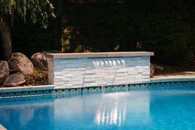 Corona F - Mt. Laurel, NJ - This custom installation features a sleek, modern waterfall constructed from white brick with a clean, geometric design. The waterfall cascades smoothly into a gunite pool, creating a tranquil water feature. The surrounding area is accented with large natural boulders, enhancing the natural aesthetic and providing a relaxing backdrop.