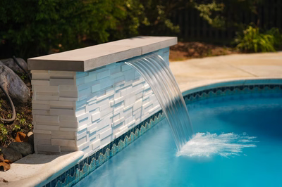 Corona H - Mt. Laurel, NJ - This professional installation features a sleek, multi-tiered stone waterfall cascading gracefully into a gunite pool. The waterfall is constructed with light-colored natural stone tiles arranged in a clean, geometric pattern, supported by a sturdy concrete coping that adds a modern touch. The pool itself showcases a vibrant blue hue and is surrounded by a well-maintained garden, enhancing the luxurious outdoor living space in Mt. Laurel, NJ.