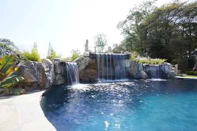 Erskine B - Colts Neck, NJ - This custom installation features a multi-tier natural stone waterfall cascading into a gunite pool, with large boulders integrated into the design for a natural, rustic look. The waterfall includes multiple spillways and a central cascade, creating a soothing water feature that flows into a serene pool area surrounded by lush landscaping and mature trees.