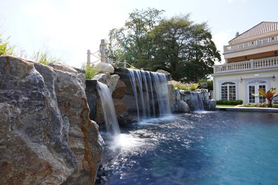 Erskine C - Colts Neck, NJ - This custom installation features a multi-tier natural stone waterfall cascading into a gunite pool, with large boulders integrated into the design for a rustic, natural look. The waterfall includes multiple spillways creating a soothing water flow, while the surrounding area is landscaped with lush greenery and a well-maintained lawn. The backdrop includes a stately home with classical architecture, enhancing the luxurious outdoor living space.