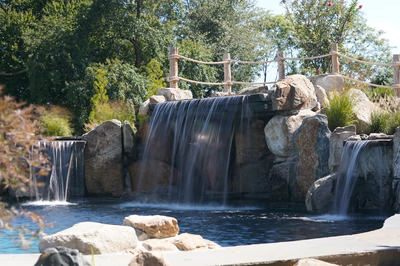 Erskine E - Colts Neck, NJ - This custom outdoor living project features a multi-tier natural stone waterfall cascading into a tranquil gunite pool. The waterfall is constructed with large, irregularly shaped boulders that create a rugged, organic look, while the water flows smoothly over each tier. The surrounding area is landscaped with lush greenery and additional boulders, enhancing the natural, serene ambiance of the Colts Neck, NJ property.