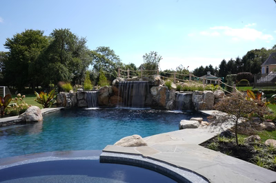 Erskine F - Colts Neck, NJ - This professionally designed outdoor living space features a multi-tier natural stone waterfall cascading into a gunite pool. The waterfall is constructed with large, irregularly shaped boulders creating a rugged, organic look, while the pool has clean lines and a smooth finish. The surrounding area is landscaped with lush greenery and decorative rock formations, enhancing the natural and serene atmosphere of the backyard setting.