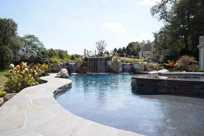 Erskine G - Colts Neck, NJ - This custom outdoor living project features a multi-tier natural stone waterfall cascading into a gunite pool with a smooth, freeform edge. The waterfall is constructed with large, irregularly shaped boulders that create a natural, cascading water flow, while the pool itself showcases a pristine, reflective surface that enhances the serene atmosphere. The surrounding area is landscaped with lush greenery and flowering plants, complemented by a stone pathway that curves gracefully around the pool, creating an inviting and tranquil backyard setting.