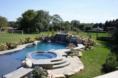 Erskine H - Colts Neck, NJ - This professionally designed outdoor living space features a multi-tier natural stone waterfall cascading into a gunite pool with a built-in spa. The waterfall is constructed from large, irregularly shaped boulders that create a rustic, freeform appearance, while the pool itself has smooth, curved edges and a deep blue water finish. The surrounding area is landscaped with manicured grass, strategically placed shrubs, and additional rock accents that enhance the natural aesthetic of the entire installation.