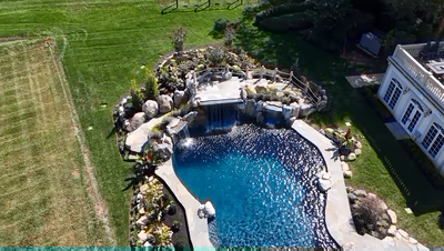 Erskine I - Colts Neck, NJ - This professionally installed outdoor feature showcases a multi-tier natural stone waterfall cascading into a gunite pool, surrounded by carefully placed boulders and lush landscaping. The waterfall's tiered design creates a soothing water flow that enhances the serene backyard setting, with the pool's smooth gunite surface reflecting sunlight beautifully. The integration of natural stone and boulders demonstrates exceptional craftsmanship and attention to detail in creating a harmonious water feature within a well-maintained garden environment.