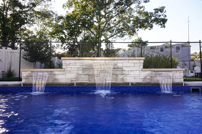 Fitzroy A - Brick, NJ - This professional installation features a multi-tier natural stone waterfall cascading into a gunite pool with blue tile accents. The waterfall is constructed from light-colored natural stone with three distinct cascades creating a soothing water feature. The pool area is surrounded by a low stone wall and features a clean, modern design with a black safety fence and lush landscaping in the background.