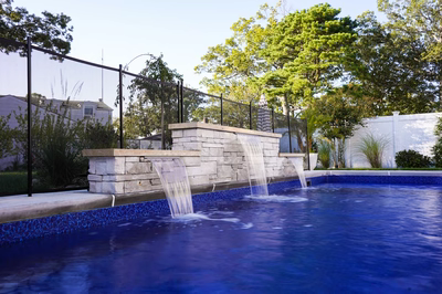 Fitzroy B - Brick, NJ - This custom installation features a multi-tier natural stone waterfall cascading gracefully into a gunite pool with vibrant blue mosaic tiles. The stone structure is meticulously crafted with clean lines and precise stonework, creating a serene water feature that enhances the backyard setting. Surrounded by lush greenery and a secure fence, the design integrates seamlessly with the residential landscape, offering both aesthetic appeal and functional relaxation.