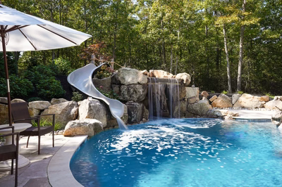 Josephine C - Mays Josephineng, NJ - This custom installation features a multi-tier natural stone waterfall with a built-in slide that cascades into a pristine gunite pool. The waterfall uses large river boulders as accents, creating a seamless, naturalistic look that blends with the surrounding woodland environment. The pool's clean lines and deep blue water contrast beautifully with the textured stonework and lush greenery, demonstrating exceptional craftsmanship and attention to detail in this backyard oasis.