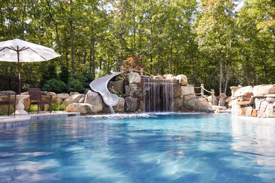 Josephine D - Mays Josephineng, NJ - This custom installation features a natural stone waterfall with a smooth, curved slide integrated into the structure, cascading water over large river rocks into a pristine gunite pool. The craftsmanship showcases tiered stonework with carefully placed boulders creating a rustic, organic feel, while the surrounding lush greenery enhances the tranquil backyard setting.