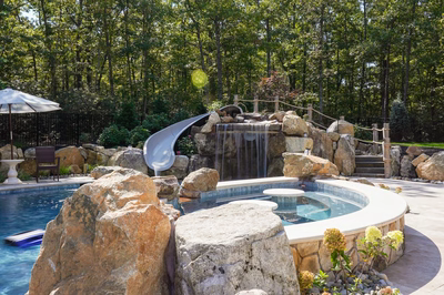 Josephine E - Mays Josephineng, NJ - This custom outdoor living project features a multi-tier natural stone waterfall cascading into a gunite pool with a built-in slide. The waterfall is constructed with large, irregularly shaped boulders creating a rugged, organic look, while the pool has a smooth, curved edge that integrates seamlessly with the surrounding landscape. The setting is enhanced by lush greenery and a wooden deck area, providing both relaxation and recreational opportunities in a serene backyard environment.