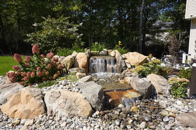 Josephine H - Mays Josephineng, NJ - This custom outdoor installation features a multi-tier natural stone waterfall cascading over large boulders into a rocky pool area surrounded by carefully arranged river stones. The waterfall includes a primary cascade and secondary spillway creating a soothing water flow, with integrated planting areas featuring vibrant flowering shrubs. The project demonstrates exceptional craftsmanship through precise stone placement and natural integration with the surrounding woodland backdrop.