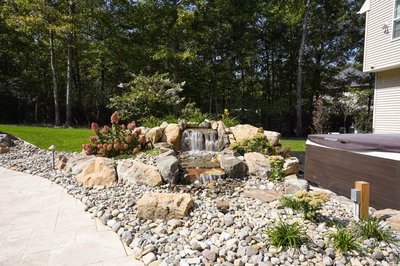 Josephine I - Mays Josephineng, NJ - This professionally installed outdoor feature showcases a multi-tier natural stone waterfall cascading over large river rocks into a rock-lined pool area. The waterfall includes a central cascade and side weirs, with carefully placed boulders creating a naturalistic grotto effect. The installation integrates seamlessly with a hot tub positioned adjacent to the pool, featuring a clean, modern design that complements the organic stone elements. The surrounding landscape includes manicured shrubs, flowering plants, and a well-maintained lawn, creating a serene backyard retreat in Mays Josephineng, NJ.