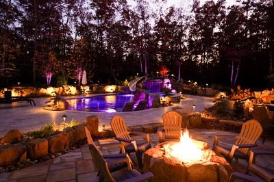 Josephine M - Mays Josephineng, NJ - This professionally designed outdoor living space features a striking natural stone waterfall cascading into a gunite pool with vibrant blue LED lighting. The installation incorporates large river boulders as accents around the pool deck and fire pit area, creating a seamless integration with the surrounding woodland environment. The multi-tiered waterfall design demonstrates exceptional craftsmanship with smooth stone surfaces and precise water flow management, while the surrounding patio includes comfortable Adirondack chairs arranged around a stone fire pit for evening relaxation.