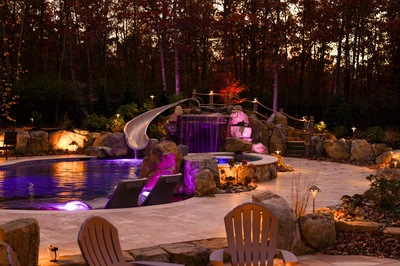 Josephine N - Mays Josephineng, NJ - This professionally designed outdoor living space features a multi-tier natural stone waterfall cascading into a gunite pool with a built-in slide. The waterfall is integrated with large boulders and illuminated with vibrant purple lighting, creating a striking visual effect. The surrounding area includes comfortable lounge chairs, a stone patio, and carefully landscaped greenery, all set against a backdrop of autumn trees, showcasing exceptional craftsmanship and attention to detail.