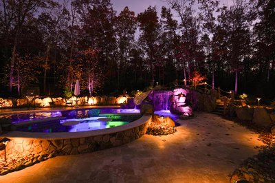 Josephine P - Mays Josephineng, NJ - This professionally designed outdoor living space features a multi-tier natural stone waterfall cascading into a gunite pool with vibrant blue LED lighting. The installation showcases exceptional craftsmanship through the seamless integration of large river boulders and carefully placed flagstone accents. The setting is enhanced by warm ambient lighting that highlights the textured stonework and creates a tranquil nighttime atmosphere in a private backyard environment.