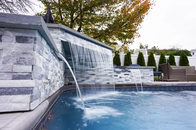 Kadishi B - Lanoka Harbor, NJ - This custom installation features a multi-tier natural stone waterfall cascading gracefully into an infinity-edge gunite pool. The waterfall is constructed with precision-cut slate and granite stones, creating a seamless, organic flow that enhances the luxurious ambiance. The surrounding area includes a well-maintained patio with wicker seating and tall, manicured evergreens, set against a backdrop of autumn foliage in Lanoka Harbor, NJ.