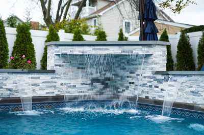 Kadishi C - Lanoka Harbor, NJ - This custom installation features a multi-tiered stone waterfall constructed from gray and white natural stone tiles, creating a sleek, contemporary design. The waterfall cascades down three levels into a gunite pool with a decorative blue tile border. The surrounding area includes neatly trimmed evergreen trees and a white fence, enhancing the luxurious backyard setting in Lanoka Harbor, NJ.