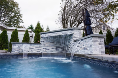 Kadishi D - Lanoka Harbor, NJ - This custom installation features a multi-tiered natural stone waterfall cascading gracefully into an infinity pool. The waterfall is constructed with carefully placed stone slabs creating a tiered cascade effect, while the pool itself showcases a sleek gunite finish with a distinctive wave-patterned coping. The surrounding area is landscaped with neatly trimmed evergreens and features a covered patio area with an umbrella, creating an inviting outdoor living space.