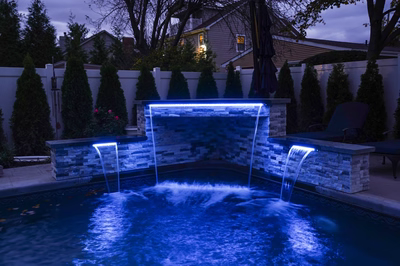 Kadishi G - Lanoka Harbor, NJ - This custom installation features a sleek, multi-tiered waterfall constructed from natural stone with integrated LED lighting that creates a striking blue glow. The waterfall cascades into a gunite pool with a smooth, reflective surface, enhancing the serene ambiance of the backyard setting. The surrounding area includes neatly trimmed conifers, a white privacy fence, and ambient outdoor lighting, showcasing high-quality craftsmanship and thoughtful design.