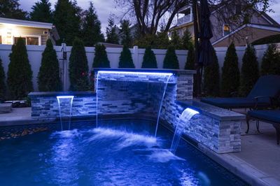 Kadishi H - Lanoka Harbor, NJ - This custom installation features a multi-tiered natural stone waterfall with integrated LED lighting that creates a striking blue glow as water cascades into a gunite pool below. The waterfall is constructed with carefully stacked stone blocks forming a clean, contemporary design that complements the surrounding manicured conifers and modern residential backdrop. The project showcases exceptional craftsmanship through precise stonework and seamless integration of water features with the pool deck.