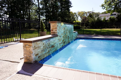 Kinsman B - Edison, NJ - This elegant outdoor living project features a custom-built natural stone waterfall cascading gracefully into a gunite swimming pool. The waterfall is constructed with layered stone tiles and a sturdy stone base, creating a soothing water feature that enhances the serene backyard setting. The pool itself has a clean, inviting blue water surface bordered by a light-colored tile deck, with a black metal fence providing privacy and security. The surrounding landscape includes well-maintained grass and mature trees, contributing to a peaceful, luxurious atmosphere perfect for relaxation and entertainment.