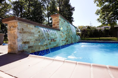 Kinsman C - Edison, NJ - This professional installation features a multi-tier natural stone waterfall cascading from a stone wall into a gunite infinity pool with vibrant blue tile accents. The waterfall includes a central spillway and smaller side cascades, showcasing exceptional craftsmanship through precise stonework and seamless integration with the pool's clean lines. The project is set in a well-maintained backyard with lush trees and a black wrought iron fence providing privacy, creating an elegant outdoor living space.