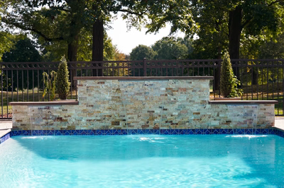 Kinsman D - Edison, NJ - This elegant outdoor installation features a multi-tiered natural stone waterfall cascading into a gunite pool. The waterfall is constructed with carefully layered stone tiles in earthy tones, creating a visually appealing water feature with multiple spillways. The pool has a clean blue surface and is bordered by a decorative blue tile edge, set within a well-maintained backyard with mature trees and a metal fence for privacy.