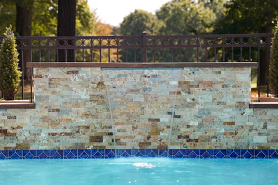 Kinsman E - Edison, NJ - This professional installation features a custom natural stone waterfall cascading gracefully into a gunite pool. The waterfall wall is constructed from a mix of beige, brown, and tan stones arranged in a staggered pattern, creating a textured and visually appealing surface. The pool deck is bordered with blue mosaic tiles, adding a vibrant contrast to the earthy tones of the stone. The surrounding area includes a wooden railing and landscaped greenery, enhancing the serene backyard setting in Edison, NJ.