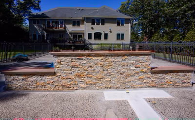 Kinsman G - Edison, NJ - This professional installation features a custom-built stone waterfall constructed from natural stone with warm-toned bricks integrated into the design. The waterfall cascades down a tiered structure with wooden seating integrated at both levels, creating an inviting outdoor living space. The project is situated in a well-maintained backyard with a modern house in the background, showcasing high-quality craftsmanship and attention to detail in both the waterfall construction and surrounding landscaping.