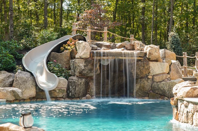 Landi A - Mays Landing, NJ - This custom outdoor installation features a multi-tier natural stone waterfall cascading into a gunite pool, with large river rocks creating a rugged, naturalistic look. A sleek, curved slide integrates seamlessly into the left side of the waterfall structure, offering both aesthetic appeal and functional recreation. The surrounding area is landscaped with lush greenery and additional boulders, enhancing the serene, woodland setting in Mays Landing, NJ.