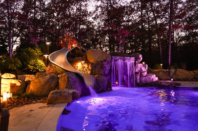 Landi J - Mays Landing, NJ - This custom outdoor living project features a striking natural stone waterfall with multiple tiers, illuminated in vibrant purple lighting that enhances the cascading water effect. The waterfall flows into a sleek gunite pool, bordered by large river rocks and integrated with a smooth concrete slide that curves gracefully from the upper tier. The setting is a well-landscaped backyard in Mays Landing, NJ, with strategic lighting highlighting the water features and surrounding foliage, creating an inviting and serene atmosphere.