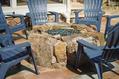 Landi L - Mays Landing, NJ - This outdoor living space features a custom-built natural stone fire pit with a central blue glass accent. The fire pit is constructed from large, irregularly shaped stones arranged in a tiered design, showcasing excellent craftsmanship and attention to detail. Surrounding the fire pit are multiple blue Adirondack chairs, creating a comfortable and inviting seating area for relaxation and social gatherings.