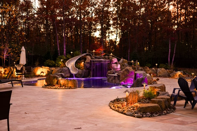 Landi O - Mays Landing, NJ - This professionally installed outdoor living project features a multi-tier natural stone waterfall cascading into a gunite pool, integrated with a smooth concrete slide for recreational use. The design incorporates large river boulders as accents around the pool deck and waterfall base, complemented by strategic LED lighting that highlights the water flow and rock textures. Set in a serene backyard environment with mature trees, the space combines natural elements with high-quality craftsmanship for a luxurious retreat.
