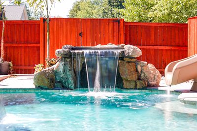 Mosquito A - DE - This custom installation features a multi-tier natural stone waterfall with smooth, flowing cascades that drop into a gunite pool below. The waterfall is constructed from large, irregularly shaped boulders and natural stone accents, creating an organic, grotto-like appearance. To the right of the waterfall, a smooth, curved slide extends from the pool deck, offering an additional recreational feature. The surrounding area includes a wooden privacy fence, potted plants, and a small house visible in the background, creating a serene backyard setting.